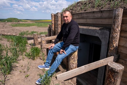 A man sits on a wooden structure in front of an earth-covered bunker with a wooden wall and doorway