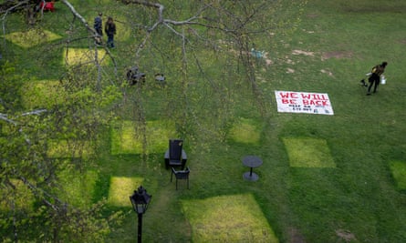 Aerial view of a green lawn with a few people walking across it, and lighter squares, with one white sign that says in read “We’ll Be Back”