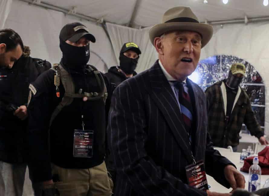 Men in military clothing and black face coverings watch over a man in a gray fedora and pinstripe suit inside a white tent.