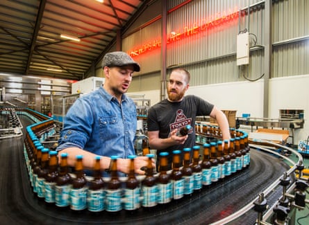 Two men stand amid a conveyor belt of bottled Punk IPA beer