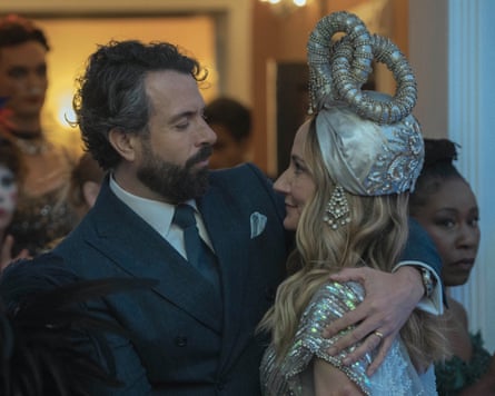 A bearded man in a suit puts his arms around a blond woman in extravagant silver costume