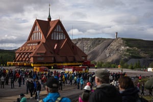A crowd watches as a wooden church sitting on yellow girders is moved on a huge flatbed truck