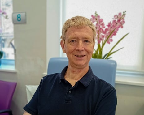 Walsh sits smiling in a dark polo shirt in a bright medical office with pink flowers behind him