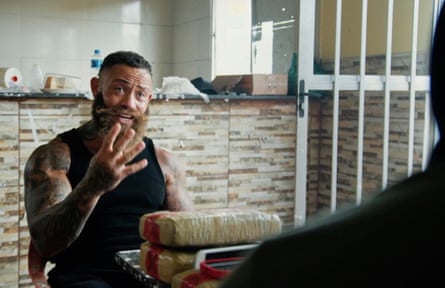Cain seated at a table with what appears to be wrapped drugs on the table in front of him, as he interviews a person for the show