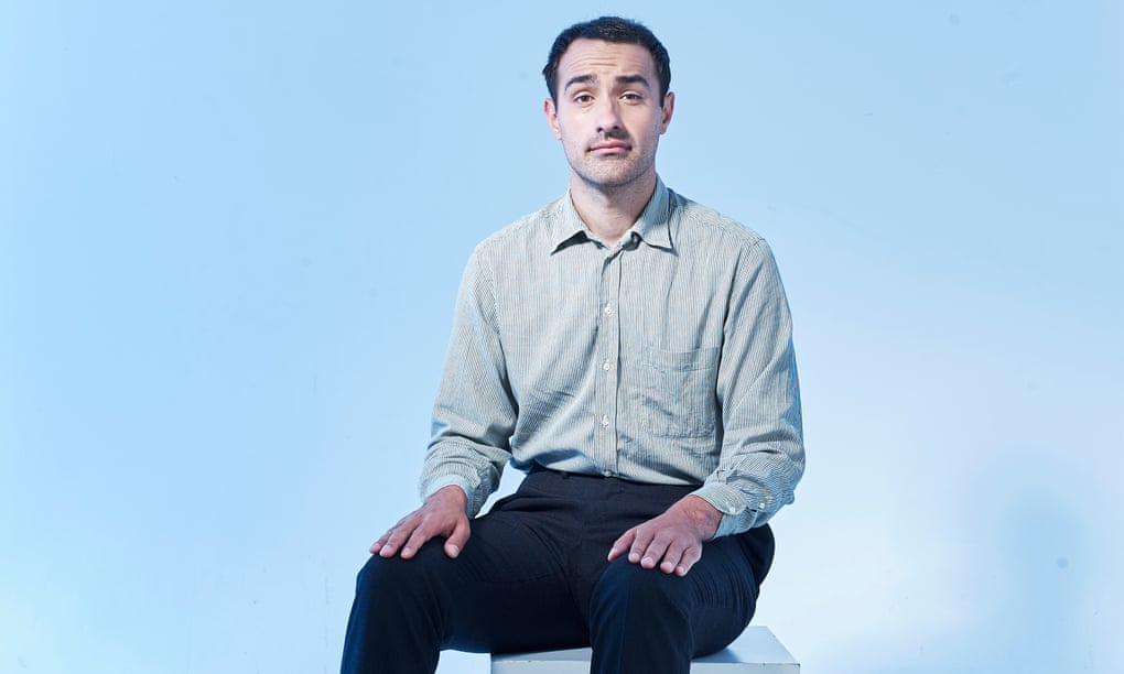 Jamie Demetriou in a pale blue shirt and black trousers, sitting on a box