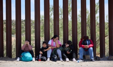 Five people, two of them children, sit in the dirt next to a tall fence made of vertical metal bars