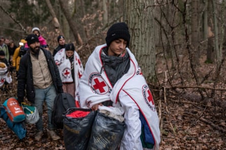 A tired family with Red Cross blankets around their shoulders walk through a forest 