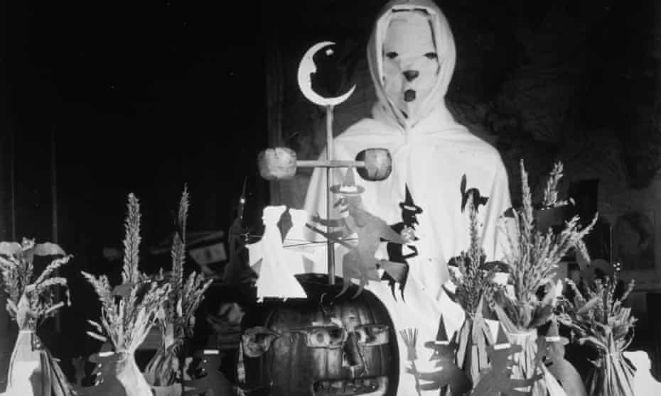 View of a person, possibly a teacher, wearing a ghost costume behind a lunch table with Halloween decorations in a rural schoolhouse. The display consists of a carved pumpkin, cutouts of witches and black cats, and haystacks, circa 1905. 