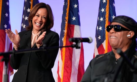 A woman gleefully claps next to a man wearing sunglasses grinning in front of a microphone