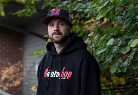 Kaylub Giamanco, a bearded man in a cap and black hoodie, stands outdoors near foliage