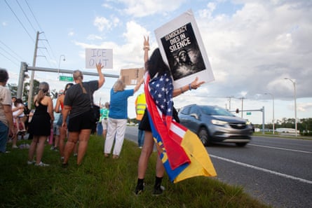 People stand in grassy area next to road as cars drive by, some holding signs with messages such as âÂÂICE outâ and âÂÂdemocracy dies in silenceâ and one wearing a flag