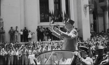 A 1958 photo of Charles de Gaulle, wearing a short flat-topped hat, gesturing at waving crowds as he stands in the back of an open car in Algeria