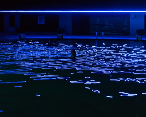 A person swims in a dark pool in Pyongyang with glowing blue light patterns projected on the surface
