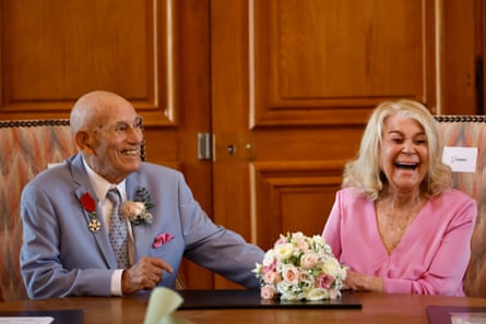 An older couple - he dressed in a power-blue suit with a medal pinned to his lapel, and she in a V-neck pink dress, sit a chairs at a glossy wooden table, in front of a bouquet of pink and white flowers, laughing.