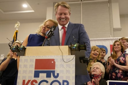 man in suit smiles while standing at lectern as a woman smiles and rests her head on his shoulder