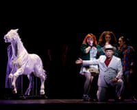 An actor kneels in a white suit beside singing performers, with a large white horse statue nearby on stage