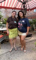 Sarah and Chloe stand smiling under a large striped garden umbrella. They have long dark hair and wear loose T-shirts over short skirts and sandals.