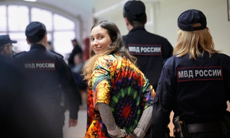 Una mujer joven con una colorida camiseta teñida sonríe por encima del hombro mientras la policía la lleva al tribunal.