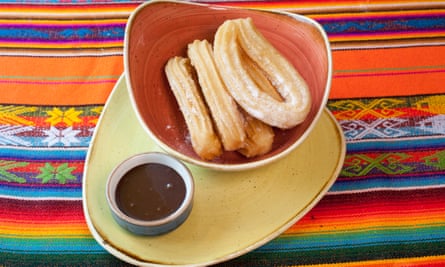 Dulce de leche churros in a red/brown bowl with a chocolate dip, both on a pale yellow plate, set on a colourful Mexican cloth