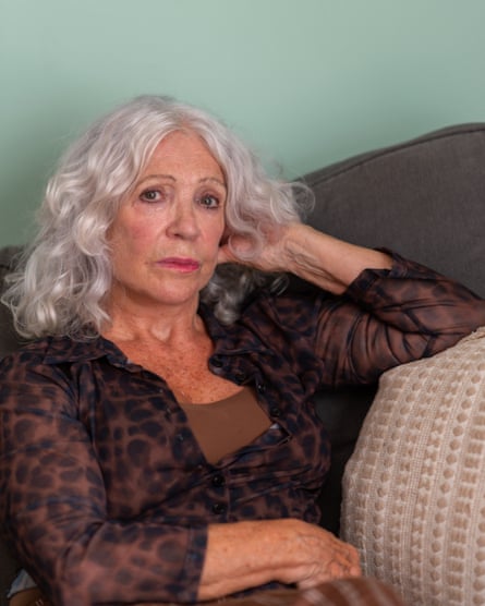 She sits on a sofa, one arm touching her neck, the other across her waist, looking at the camera