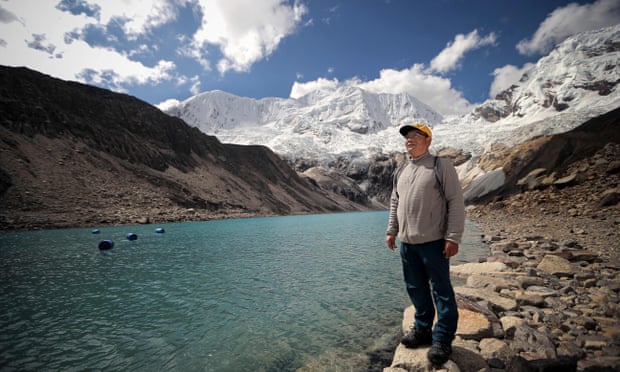 A man stands on a rocky shore next to a clear blue lake with glacial mountains in the background.