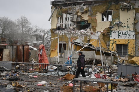 A man walks through debris with a heavily damaged building seen in the background.