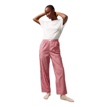 A model wears a white T-shirt tucked into pink and red striped poplin pyjamas and oatmeal coloured socks. The model is a Black person with their hands behind their head and very short hair.