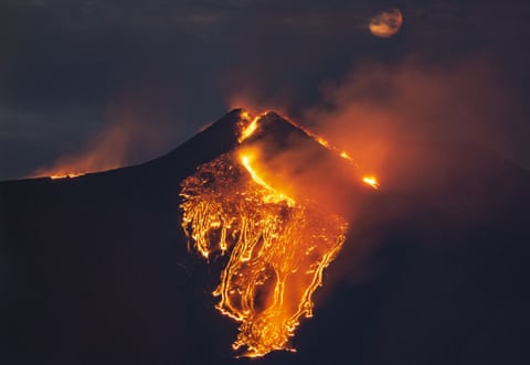The moon is partially seen in the sky as lava flows from the Mount Etna volcano