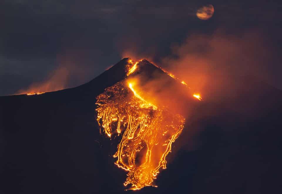 The moon is partially seen in the sky as lava flows from the Mount Etna volcano
