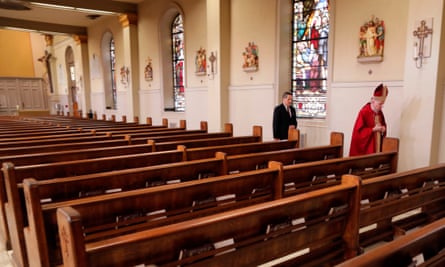 Inside of a church with bare pews, and an older man with a red pointy hat and red robe, followed by another man in a dark suit, alongside stained-glass windows.