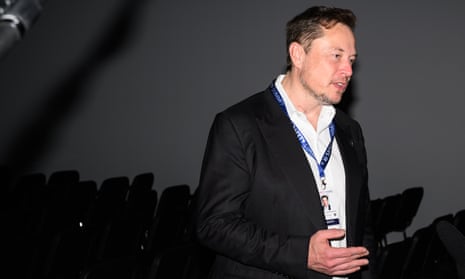 Elon Musk speaks with members of the media – he wears white shirt with open neck, a black jacket and a lanyard with security pass