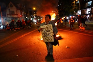 A woman holds a poster that reads ‘Alvaro Uribe goes to prison’. Behind her are fires and other people gathered on the street