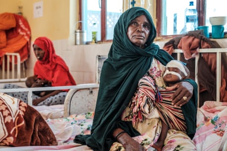 Two African women with cloaks wrapped round their head and shoulders sit on hospital ward holding malnourished babies