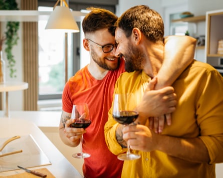 Happy hipster men in love hugging while standing in the kitchen. They are cooking dinner together at home and drinking wine