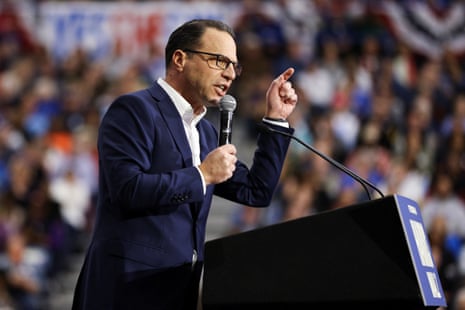 a man in a suit points while speaking into a microphone from behind a lectern to a crowd of people