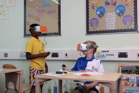 Two boys wear what appear to be VR masks in a classroom; one boy, who wears a yellow polo shirt and colourful patterned shorts, is standing and the other, in a blue and white sports top, sits at a desk. One boy is black and the other white with pale skin and fair hair.