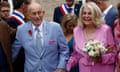 Older white couple, he in powder-blue suit with pink kerchief and red and war medal on lapel, she in V-necked pink dress and holding white and pink bouquet, hold hands, grinning, as they walk through a crowd.