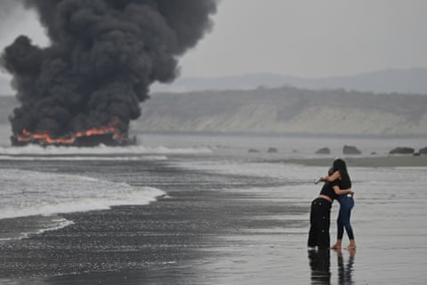 Two young women hug as they stand on a wet beach beside lapping waves. Thick black smoke is rising from the flames as boats burn further down the beach