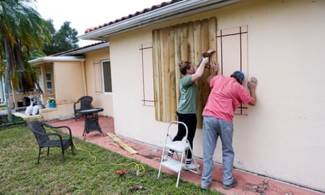 person wearing green shirt uses drill to board up home as another person wearing pink shirt presses against board
