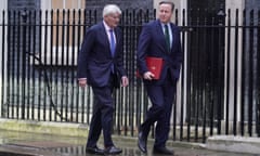 Two men in suits walk along a pavement next to high black railings, one of them holding a red folder