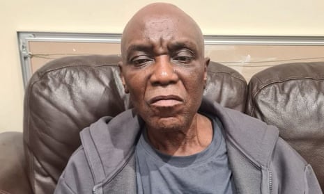 A Nigerian 61-year-old man looks is sitting on a brown leather sofa and looking straight ahead into the camera