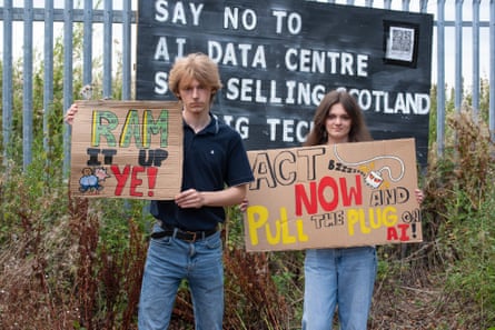 A young man and women hold handpainted cardboard signs and stand in front of a black banner that reads ‘Say no to AI Data Centre’ which is fastened on a metal fence. Their signs read ‘Ram it up ye!’ and ‘Act now, pull the plug on AI’.
