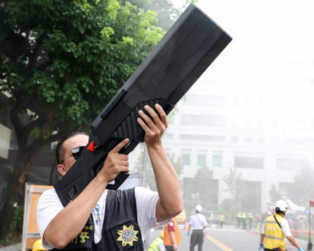 A man standing in a city street points an anti-drone weapon into the sky. It is shaped like a rifle but with a very wide gun barrel
