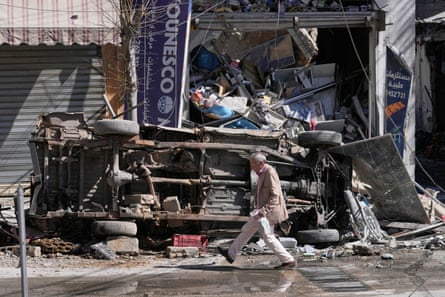 A man walks past the mangled wreckage of a car laying on its side, and a destroyed shop