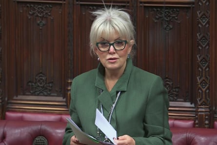 she has her white-blond hair tied up with a fringe over her forehead, wears dark-rimmed glasses and a dark green jacket.