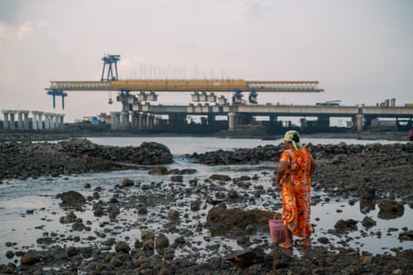 A woman with a basket standing on a rocky foreshore with a highway on piles being built on the horizon