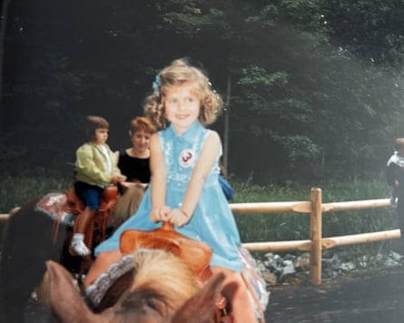Bauer as a girl wearing a blue dress with a badge with the number 3 riding a horse and smiling.
