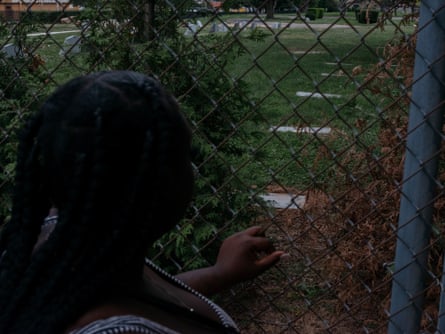 Seen from behind, a young Black woman with long black braids, holds onto a chainlink fence and looks toward stone grave markers in grass on the other side.