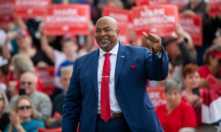 Large bald Black man in blue suit and red tie, smiling and pumping fists, with wave of people holding up red signs behind him.