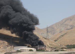 A thick cloud of black smoke rises from an oil storage facility in a desert valley